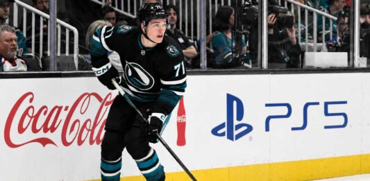 Macklin Celebrini Point Projections 2026-27: San Jose Sharks Fantasy Hockey Outlook Macklin Celebrini skating for the San Jose Sharks with Coca-Cola and PS5 board ads, highlighting his 2026-27 fantasy hockey point projection