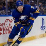 Tage Thompson Point Projections 2026-27: Buffalo Sabres Fantasy Hockey Outlook Tage Thompson skating right-handed along the boards in a Buffalo Sabres jersey beside Coca-Cola and PS5 ads during an NHL game.