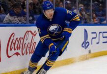 Tage Thompson Point Projections 2026-27: Buffalo Sabres Fantasy Hockey Outlook Tage Thompson skating right-handed along the boards in a Buffalo Sabres jersey beside Coca-Cola and PS5 ads during an NHL game.