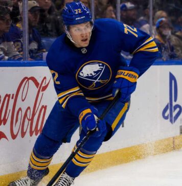 Tage Thompson Point Projections 2026-27: Buffalo Sabres Fantasy Hockey Outlook Tage Thompson skating right-handed along the boards in a Buffalo Sabres jersey beside Coca-Cola and PS5 ads during an NHL game.