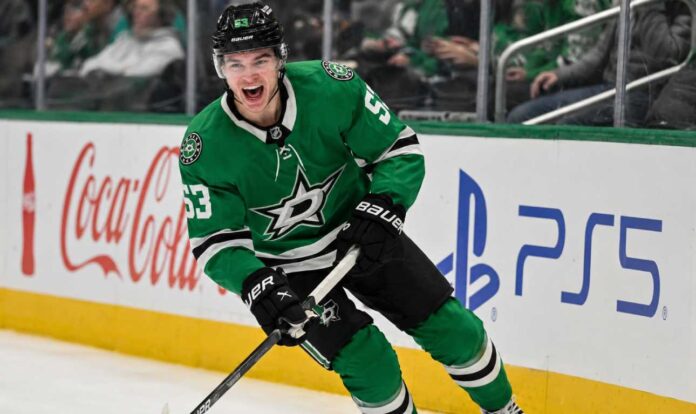 wyatt-johnston-dallas-stars-fantasy-hockey-projection-2026-27 Wyatt Johnston skating in a Dallas Stars home jersey beside Coca-Cola and PS5 ads for his 2026-27 fantasy hockey point projection.