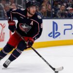 Zach Werenski skating left-handed for the Columbus Blue Jackets beside Coca-Cola and PS5 board ads during a 2026-27 fantasy hockey projection