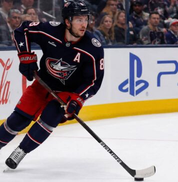 Zach Werenski skating left-handed for the Columbus Blue Jackets beside Coca-Cola and PS5 board ads during a 2026-27 fantasy hockey projection