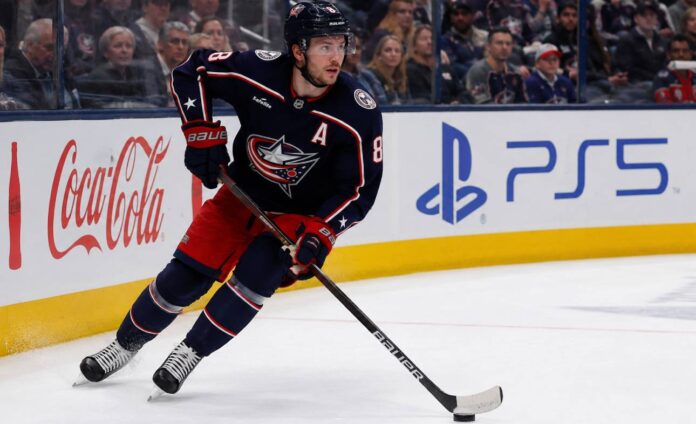 Zach Werenski skating left-handed for the Columbus Blue Jackets beside Coca-Cola and PS5 board ads during a 2026-27 fantasy hockey projection