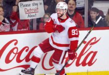 Alex DeBrincat celebrates for the Detroit Red Wings as a fan holds a “Please stay in Detroit Alex!” sign amid NHL trade rumors.