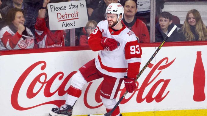 alex-debrincat-detroit-red-wings-trade-rumors Alex DeBrincat celebrates for the Detroit Red Wings as a fan holds a “Please stay in Detroit Alex!” sign amid NHL trade rumors.