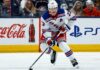 Adam Fox Point Projections 2026-27: New York Rangers Fantasy Outlook Adam Fox skating for the New York Rangers beside Coca-Cola and PS5 board ads in a 2026-27 fantasy hockey point projection.