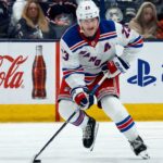 Adam Fox Point Projections 2026-27: New York Rangers Fantasy Outlook Adam Fox skating for the New York Rangers beside Coca-Cola and PS5 board ads in a 2026-27 fantasy hockey point projection.