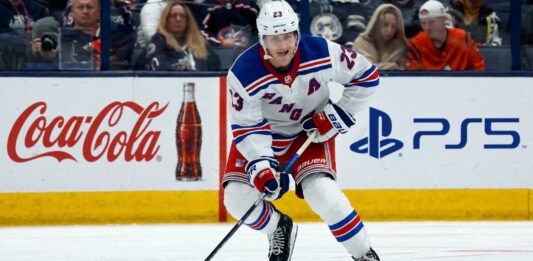 Adam Fox Point Projections 2026-27: New York Rangers Fantasy Outlook Adam Fox skating for the New York Rangers beside Coca-Cola and PS5 board ads in a 2026-27 fantasy hockey point projection.