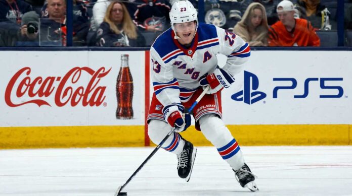 adam-fox-new-york-rangers-fantasy-hockey-projection-2026-27 Adam Fox skating for the New York Rangers beside Coca-Cola and PS5 board ads in a 2026-27 fantasy hockey point projection.