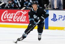 Dylan Guenther Point Projections 2026-27: Utah Mammoth Fantasy Outlook Dylan Guenther skating for the Utah Mammoth beside Coca-Cola and PS5 board ads in a 2026-27 fantasy hockey point projection.