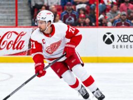 Dylan Larkin skating for the Detroit Red Wings amid NHL trade rumors, with Colorado Avalanche fans in the stands and Coca-Cola and Xbox Series X ads on the boards.