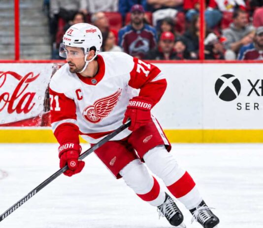 Dylan Larkin skating for the Detroit Red Wings amid NHL trade rumors, with Colorado Avalanche fans in the stands and Coca-Cola and Xbox Series X ads on the boards.