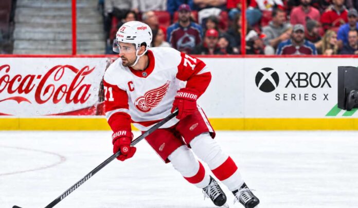 dylan-larkin-detroit-red-wings-trade-rumors-2026 (1) Dylan Larkin skating for the Detroit Red Wings amid NHL trade rumors, with Colorado Avalanche fans in the stands and Coca-Cola and Xbox Series X ads on the boards.