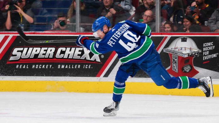 elias-pettersson-vancouver-canucks-los-angeles-kings-trade-rumors Elias Pettersson of the Vancouver Canucks takes a powerful shot in action as a Super Chexx bubble hockey board ad appears behind him amid Los Angeles Kings trade rumors.