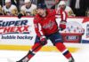Jordan Kyrou in a Washington Capitals home jersey skates with the puck in front of a stylish Super Chexx bubble hockey boards ad and Pittsburgh Penguins bench