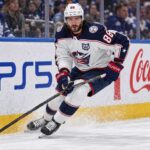 Montreal Canadiens Trade Rumors: Could Kirill Marchenko Be Traded Before Opening Night? Kirill Marchenko skating for the Columbus Blue Jackets beside PS5 and Coca-Cola board ads amid Montreal Canadiens trade rumors.