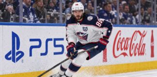 Montreal Canadiens Trade Rumors: Could Kirill Marchenko Be Traded Before Opening Night? Kirill Marchenko skating for the Columbus Blue Jackets beside PS5 and Coca-Cola board ads amid Montreal Canadiens trade rumors.