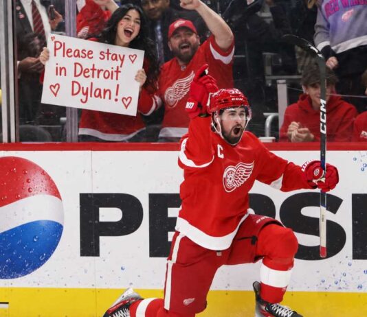 Detroit Red Wings captain Dylan Larkin celebrates on one knee in front of cheering fans, including a female fan holding a “Please stay in Detroit Dylan!” sign, with a Pepsi board ad in the background.