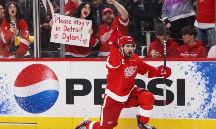 nhl-news-rumors-september-26-2026 Detroit Red Wings captain Dylan Larkin celebrates on one knee in front of cheering fans, including a female fan holding a “Please stay in Detroit Dylan!” sign, with a Pepsi board ad in the background.