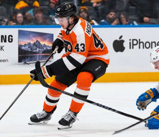 Quinn Hughes Trade Rumors: Could the Philadelphia Flyers Beat the Devils to the Wild Superstar? Quinn Hughes in a Philadelphia Flyers home uniform during NHL trade rumor speculation, with Samsung TV and Apple iPhone rink board ads in the background