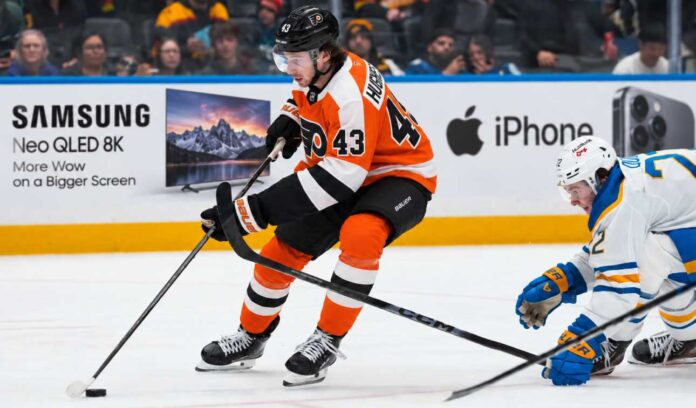 Quinn Hughes in a Philadelphia Flyers home uniform during NHL trade rumor speculation, with Samsung TV and Apple iPhone rink board ads in the background