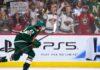 Quinn Hughes Trade Rumors: Are the Wild Running Out of Time Before a Devils Blockbuster? Minnesota Wild defenseman Quinn Hughes skates in front of a PS5 rink-board ad as a New Jersey Devils fan behind the glass holds a sign reading “Come to New Jersey Quinn!” during NHL trade rumor buzz.
