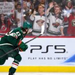 Quinn Hughes Trade Rumors: Are the Wild Running Out of Time Before a Devils Blockbuster? Minnesota Wild defenseman Quinn Hughes skates in front of a PS5 rink-board ad as a New Jersey Devils fan behind the glass holds a sign reading “Come to New Jersey Quinn!” during NHL trade rumor buzz.
