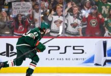 Quinn Hughes Trade Rumors: Are the Wild Running Out of Time Before a Devils Blockbuster? Minnesota Wild defenseman Quinn Hughes skates in front of a PS5 rink-board ad as a New Jersey Devils fan behind the glass holds a sign reading “Come to New Jersey Quinn!” during NHL trade rumor buzz.