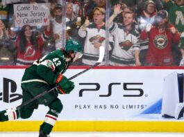 Quinn Hughes Trade Rumors: Are the Wild Running Out of Time Before a Devils Blockbuster? Minnesota Wild defenseman Quinn Hughes skates in front of a PS5 rink-board ad as a New Jersey Devils fan behind the glass holds a sign reading “Come to New Jersey Quinn!” during NHL trade rumor buzz.