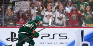Quinn Hughes Trade Rumors: Are the Wild Running Out of Time Before a Devils Blockbuster? Minnesota Wild defenseman Quinn Hughes skates in front of a PS5 rink-board ad as a New Jersey Devils fan behind the glass holds a sign reading โCome to New Jersey Quinn!โ during NHL trade rumor buzz.