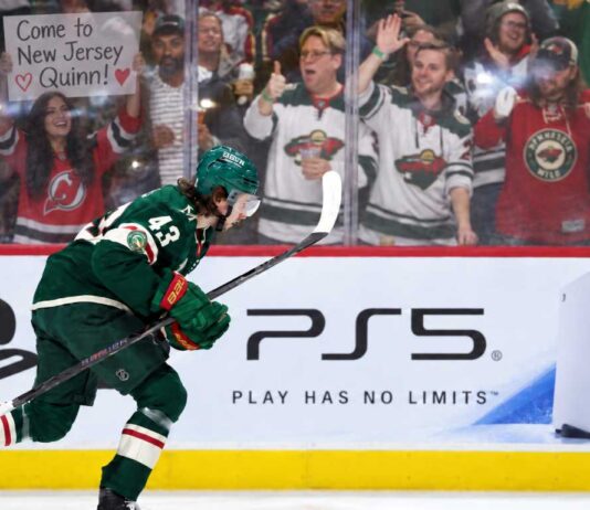 Quinn Hughes Trade Rumors: Are the Wild Running Out of Time Before a Devils Blockbuster? Minnesota Wild defenseman Quinn Hughes skates in front of a PS5 rink-board ad as a New Jersey Devils fan behind the glass holds a sign reading “Come to New Jersey Quinn!” during NHL trade rumor buzz.