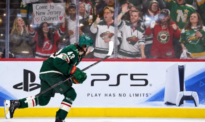 quinn-hughes-wild-devils-trade-rumors-sept-2026 Minnesota Wild defenseman Quinn Hughes skates in front of a PS5 rink-board ad as a New Jersey Devils fan behind the glass holds a sign reading “Come to New Jersey Quinn!” during NHL trade rumor buzz.