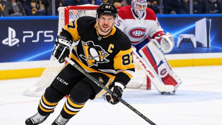 Sidney Crosby skating in a Pittsburgh Penguins home jersey with a Montreal Canadiens goalie in the background and a PS5 rink-board advertisement.