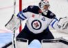 Connor Hellebuyck of the Winnipeg Jets in his white away jersey making a save as NHL trade rumors link the star goaltender to the Buffalo Sabres and Utah Mammoth ahead of the October 23 decision.