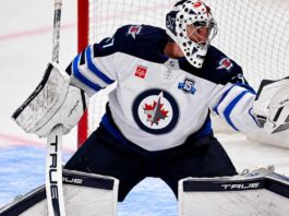 Connor Hellebuyck of the Winnipeg Jets in his white away jersey making a save as NHL trade rumors link the star goaltender to the Buffalo Sabres and Utah Mammoth ahead of the October 23 decision.