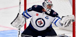 Connor Hellebuyck of the Winnipeg Jets in his white away jersey making a save as NHL trade rumors link the star goaltender to the Buffalo Sabres and Utah Mammoth ahead of the October 23 decision.