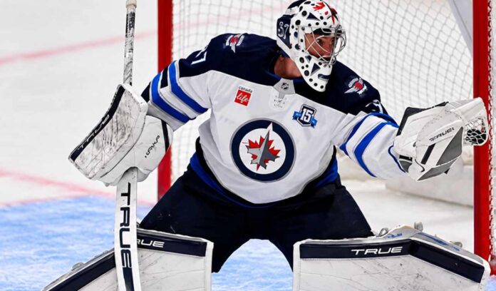 Connor Hellebuyck of the Winnipeg Jets in his white away jersey making a save as NHL trade rumors link the star goaltender to the Buffalo Sabres and Utah Mammoth ahead of the October 23 decision.