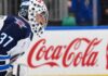 Winnipeg Jets goaltender Connor Hellebuyck kneeling on the ice in his white away jersey with a Coca-Cola rink board in the background. The suspended goalie is forfeiting over $38,000 per day in lost salary.