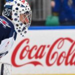 Winnipeg Jets goaltender Connor Hellebuyck kneeling on the ice in his white away jersey with a Coca-Cola rink board in the background. The suspended goalie is forfeiting over $38,000 per day in lost salary.