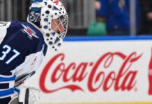 Winnipeg Jets goaltender Connor Hellebuyck kneeling on the ice in his white away jersey with a Coca-Cola rink board in the background. The suspended goalie is forfeiting over $38,000 per day in lost salary.