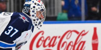 Winnipeg Jets goaltender Connor Hellebuyck kneeling on the ice in his white away jersey with a Coca-Cola rink board in the background. The suspended goalie is forfeiting over $38,000 per day in lost salary.