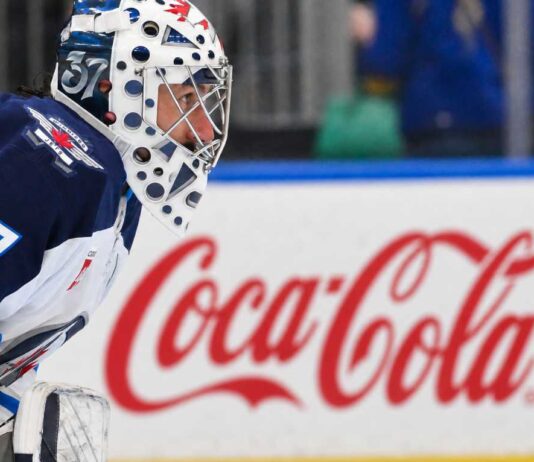 Winnipeg Jets goaltender Connor Hellebuyck kneeling on the ice in his white away jersey with a Coca-Cola rink board in the background. The suspended goalie is forfeiting over $38,000 per day in lost salary.