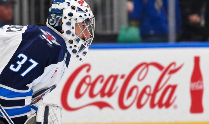 connor-hellebuyck-salary-loss-tracker Winnipeg Jets goaltender Connor Hellebuyck kneeling on the ice in his white away jersey with a Coca-Cola rink board in the background. The suspended goalie is forfeiting over $38,000 per day in lost salary.