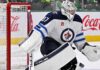 Did the Winnipeg Jets Just Make a Connor Hellebuyck Trade More Expensive? Connor Hellebuyck in Winnipeg Jets goalie equipment defending the net with a Coca-Cola advertisement on the boards amid NHL trade rumors.