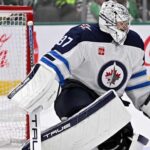 Did the Winnipeg Jets Just Make a Connor Hellebuyck Trade More Expensive? Connor Hellebuyck in Winnipeg Jets goalie equipment defending the net with a Coca-Cola advertisement on the boards amid NHL trade rumors.