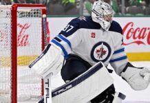 Did the Winnipeg Jets Just Make a Connor Hellebuyck Trade More Expensive? Connor Hellebuyck in Winnipeg Jets goalie equipment defending the net with a Coca-Cola advertisement on the boards amid NHL trade rumors.
