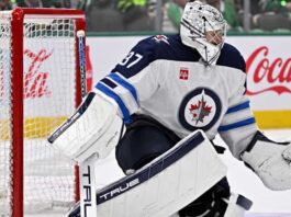 Connor Hellebuyck in Winnipeg Jets goalie equipment defending the net with a Coca-Cola advertisement on the boards amid NHL trade rumors.