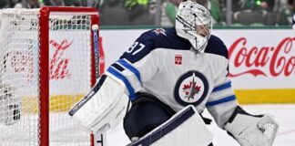 Connor Hellebuyck in Winnipeg Jets goalie equipment defending the net with a Coca-Cola advertisement on the boards amid NHL trade rumors.