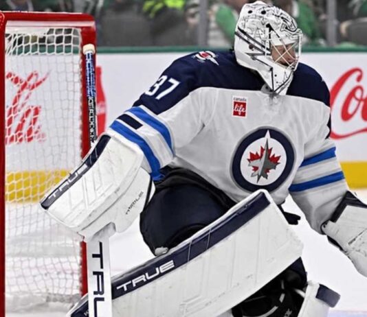 Connor Hellebuyck in Winnipeg Jets goalie equipment defending the net with a Coca-Cola advertisement on the boards amid NHL trade rumors.
